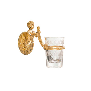 This toothbrush holder features a smooth and elegant cylindrical body made of cut crystal, with faceted edges that add a tactile dimension and reflect light beautifully. The base is crafted from polished brass, providing a sturdy and cool-to-the-touch foundation. A detailed cherub motif is intricately sculpted into the brass, offering a raised and textured design that adds a classical and artistic feel.